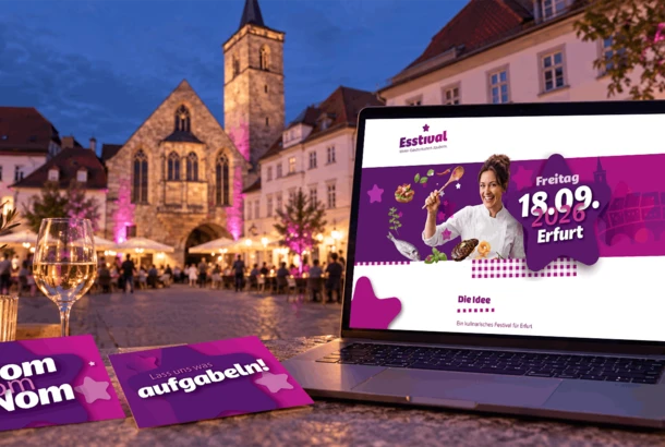 Laptop mit Esstival-Erfurt-Website und zwei lila Postkarten vor dem abendlich beleuchteten Wenigemarkt in Erfurt mit pink angestrahlten Bäumen.