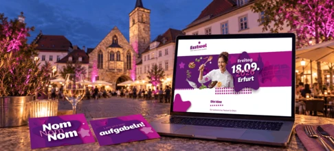 Laptop mit Esstival-Erfurt-Website und zwei lila Postkarten vor dem abendlich beleuchteten Wenigemarkt in Erfurt mit pink angestrahlten Bäumen.