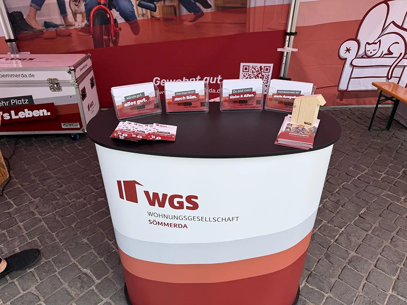 Gebrandete WGS-Infotheke mit Postkarten, Flyern, QR-Code und kleinen Holzhaus-Giveaways.
